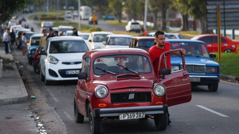 A man in an orange t-shirt pushes his old-model Fiat car with the door open, with two lanes of cars lined up behind him