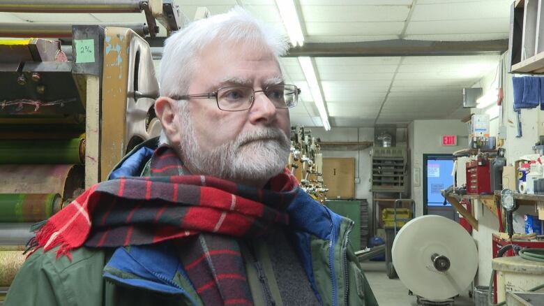 A man in a beard and a scarf stands in a room in front of a printing press.