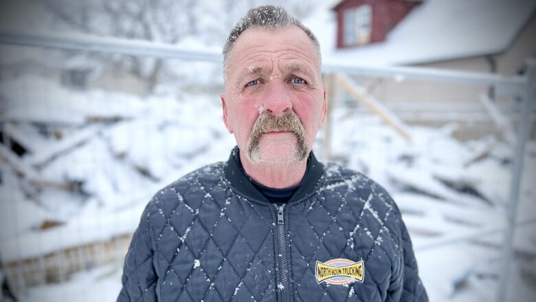 A man standing in front of a demolished home looks at the camera as snow falls.