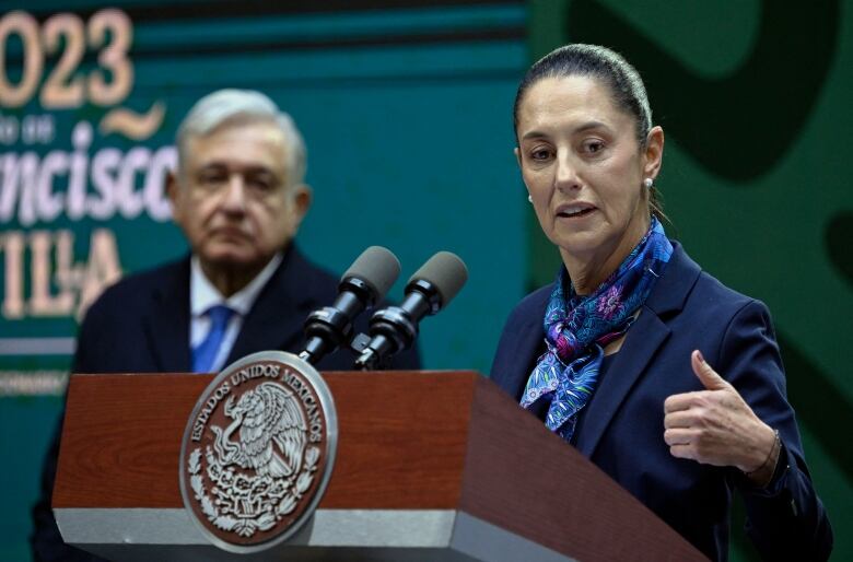 A woman with her hair pulled back wearing a scarf speaks at a podium as an older man in a suit and tie looks on from the side.