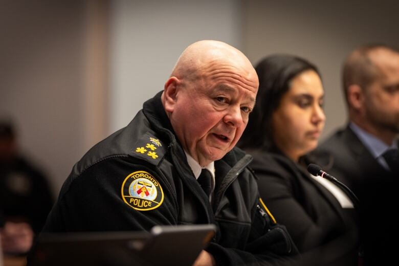 Toronto's police chief, wearing a back jacket with police logo on it, speaks while seated at a city committee table.