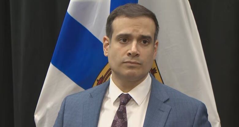 A man in a suit stands in front of a Nova Scotia flag.