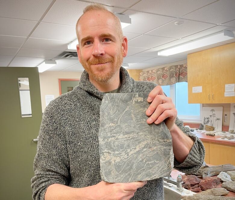 A young man with reddish hair, beard and moustache and a grey sweater smiles and holds up a grey rock containing fossils.