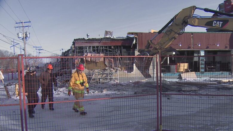 three people, one in a firefighter suit, walk in a gated area with a building partially wrecked in the background