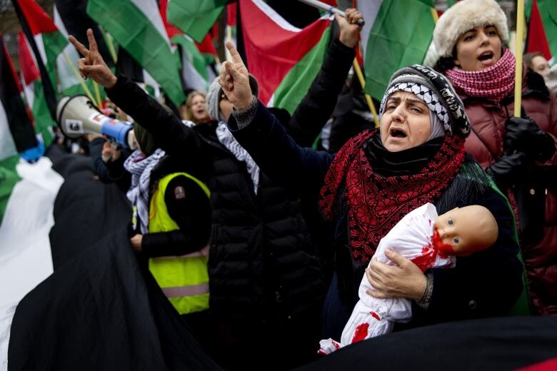 Several people are shown raising arms and chanting while holding flags and signs at an outdoor protest. One woman in a headscarf holds a baby doll with what appears to be red paint on it.