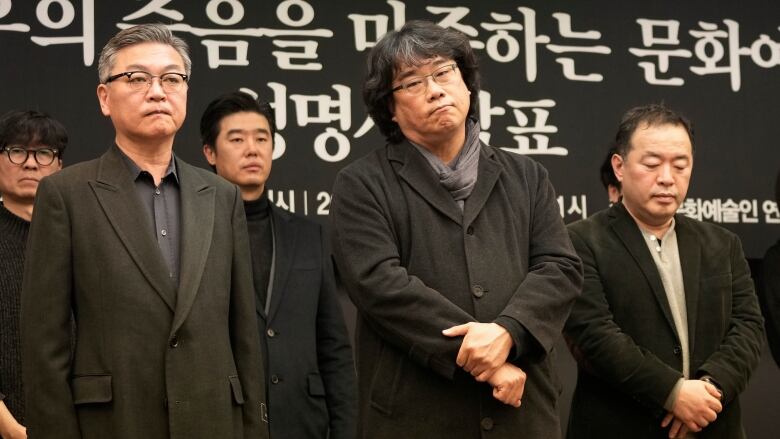 Several men stand solemnly in front of a black backdrop with words written in Korean.