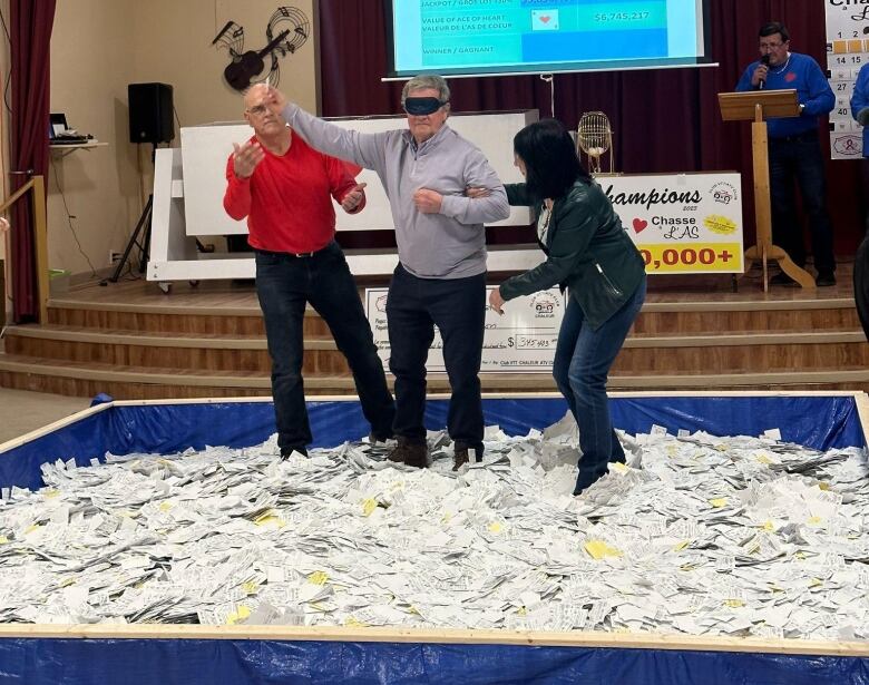 A man wades through a sea of papers while blindfolded, holding a piece of paper up in his right hand.