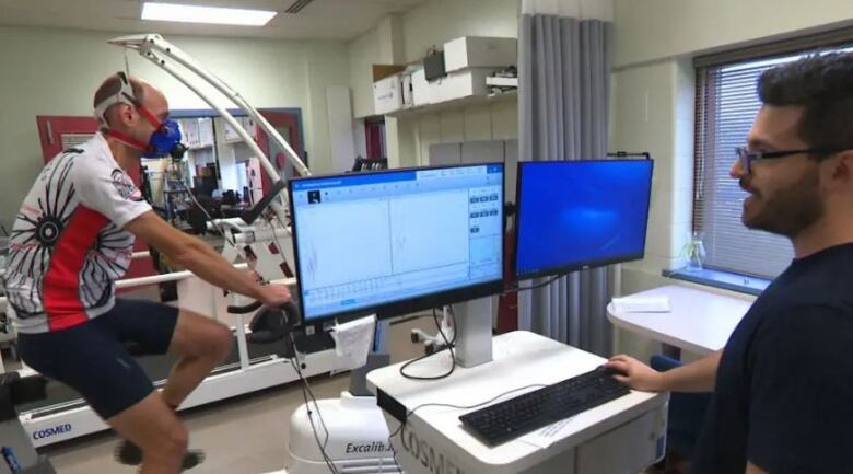 Man on bike wearing a mask cycles in a lab. A technician watches results come up on computer screens.