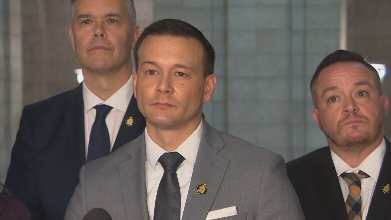 A man in a suit stands at a microphone. Two other men in suits stand behind him.