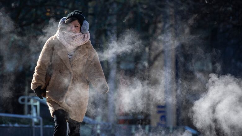 A woman walks while wearing a coat and earmuffs, as steam emanates around her.