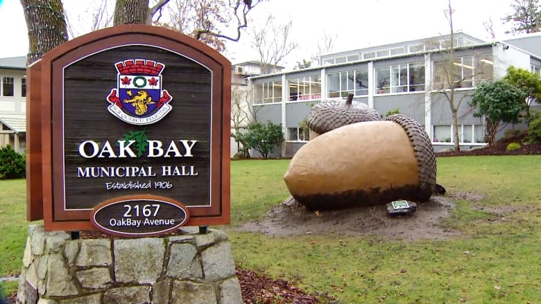 A sign reading 'Oak Bay Municipal Hall' is pictured on the foreground. In the background, a large sculpture depicting two acorns nestling against each other is seen in front of a building. 