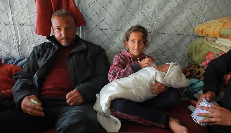 A man sits with a girl who is holding a baby.