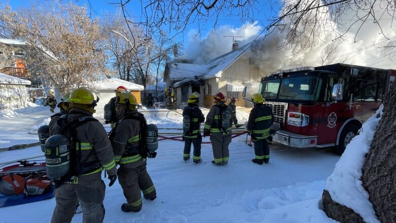 Firefighters stand on a snowy road with a fire truck behind them, and a house with smoke billowing out of it behind that.