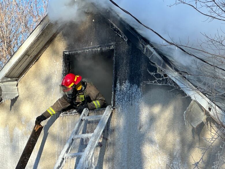 A firefighter leans out of an upper-story window, with smoke visible behind him, and appears to be removing a burnt piece of wood from the building.