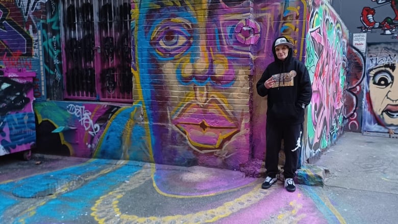 A man wearing a black hoodie poses in front of a purple-pink mural, and is surrounded by numerous others in an alley.