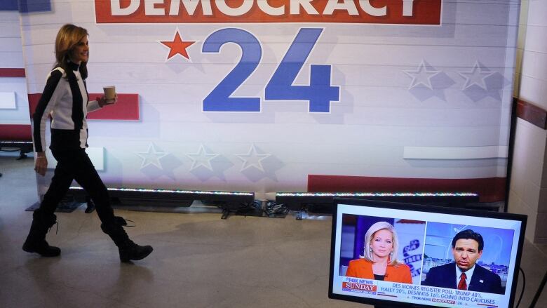 Nikki Haley walks on a stage; in the foreground there's a TV with Ron DeSantis on it 