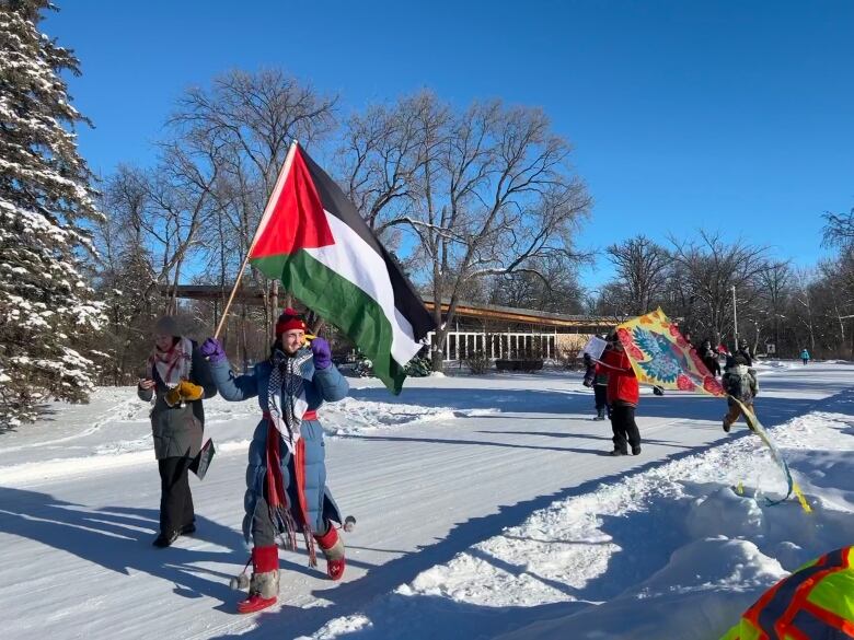 A person dressed in winter clothing walks down a snowy road holding the flag of Palestine.