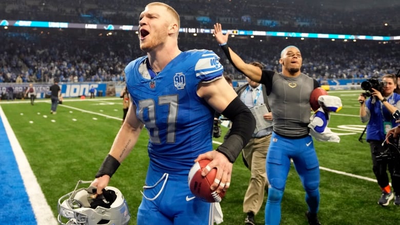 A male football player wearing number 97 shouts in celebration while holding a ball in his left hand as a teammate raises their right arm while following behind him off the field.