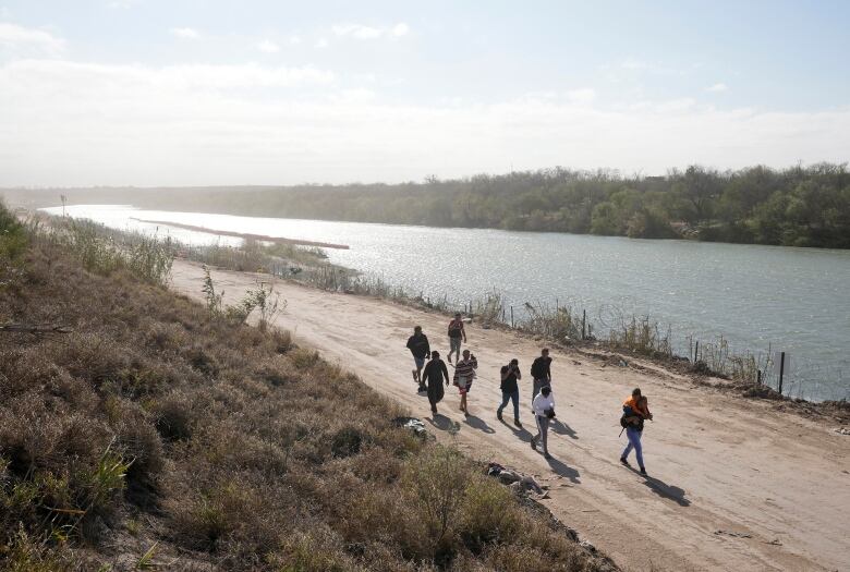 Several people are shown from a distance walking along a dirt pass beside a body of water.