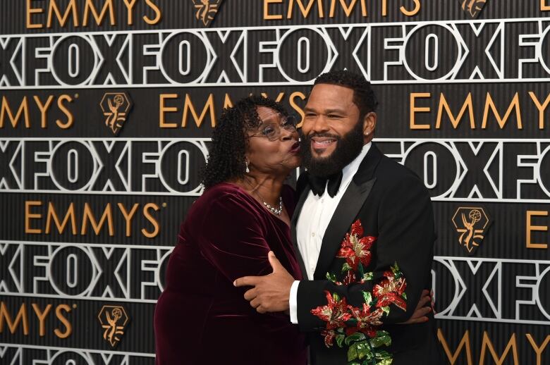 A Black woman in a wine-coloured velvet gown kisses the cheek of a Black man in a tuxedo jacket with a flower applique. 