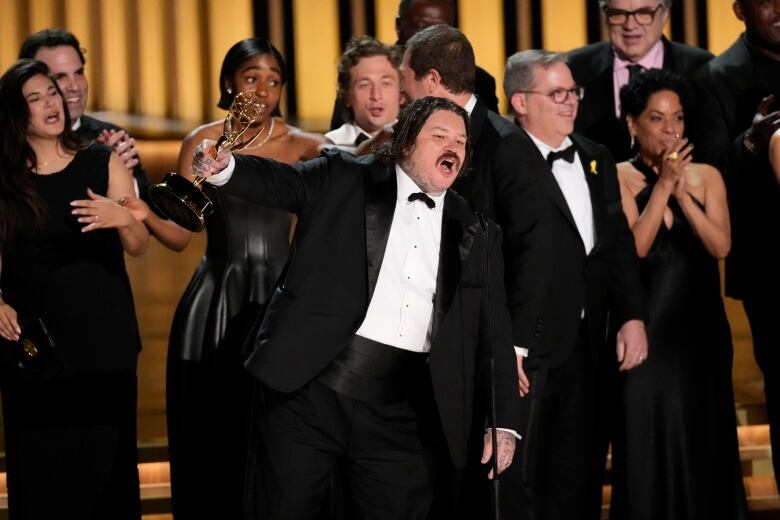 A man in a tuxedo holds an Emmy Award while speaking on stage. 