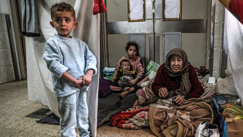 An elderly woman and three young children sit in a makeshift shelter, made of hanging sheets, inside a hospital in Gaza.