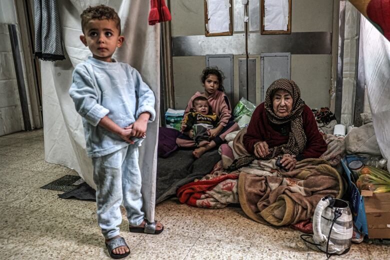 An elderly woman and three young children sit in a makeshift shelter, made of hanging sheets, inside a hospital in Gaza.