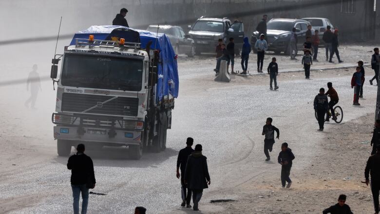 A truck with a man riding atop it is shown travelling down a dirt road as dozens of onlookers are shown at each side of the road.