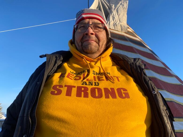 A man in a yellow hoodie and a black jacket stands in front of a sweat lodge. The hoodie bears the message 