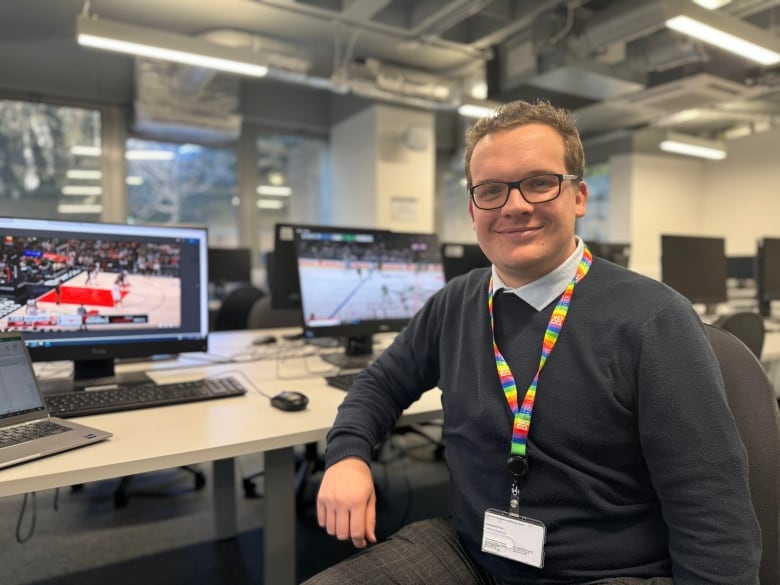 A man poses in a chair looking at the camera and smiling. He has computer monitors behind him with footage from a basketball game and a hockey game.