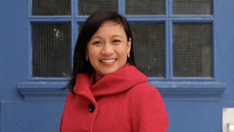 Filipino woman in red coat with long black hair standing in front of blue wall with window.