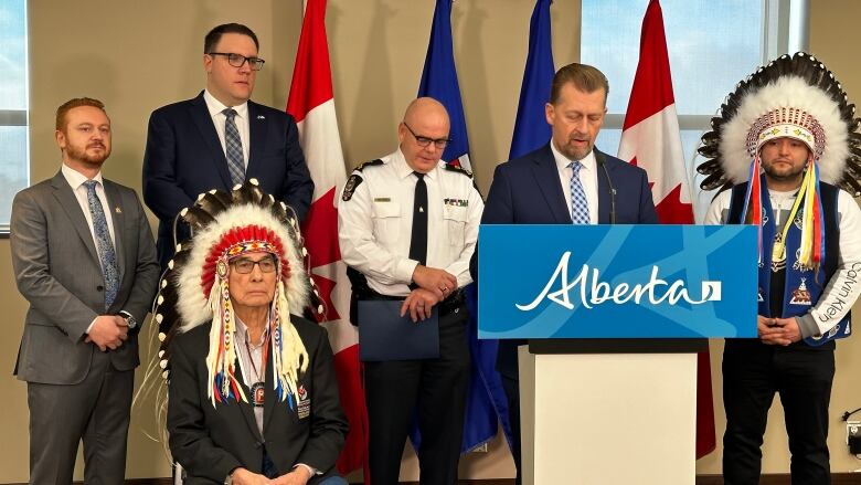 Six men in suits, a police uniform, and head gear stand on a podium. 