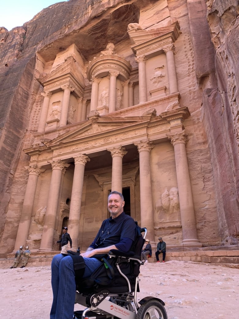 A man in an electric wheelchair smiles widely while posing in front of an ancient building while travelling. 