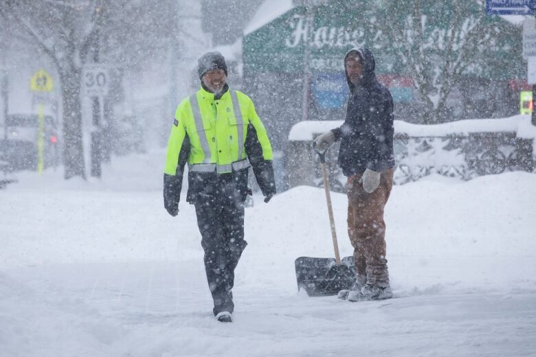 A male Canada Post worker walks past a man shoveling sidewalk snow.