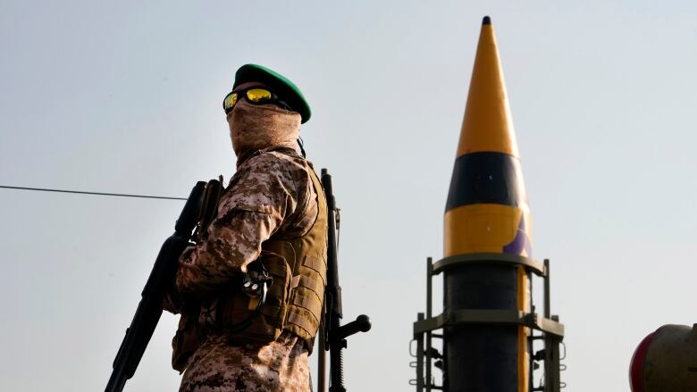 A man in military garb carrying guns stands in the foreground. Behind is a missilehead.