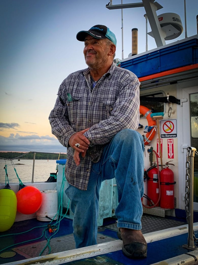 A fisher wearing a plaid shirt stands on his fishing boat with one foot on the gunwale, looking out to sea. 