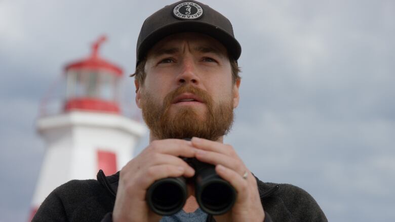 A man with a beard and wearing a hat holds binoculars in his hand and looks out beyond camera. 