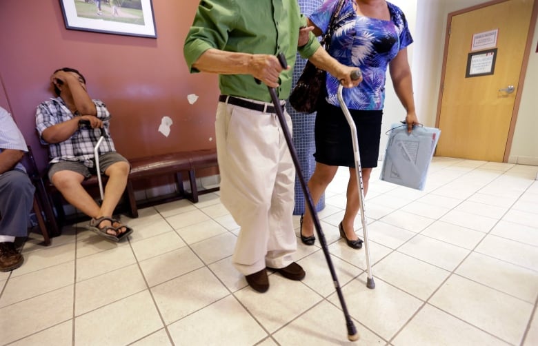 A man using two canes is helped in the waiting area of a doctor's office.