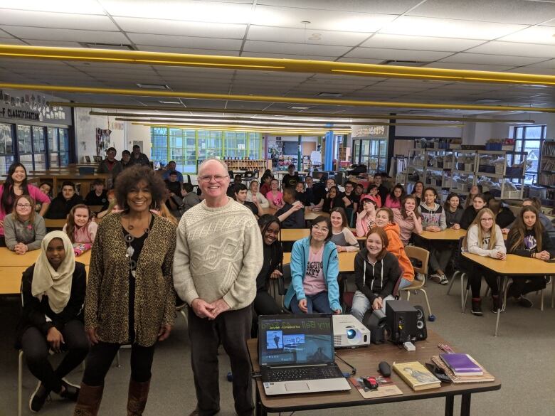 A Black woman and white man stand in front of children in a school library. 