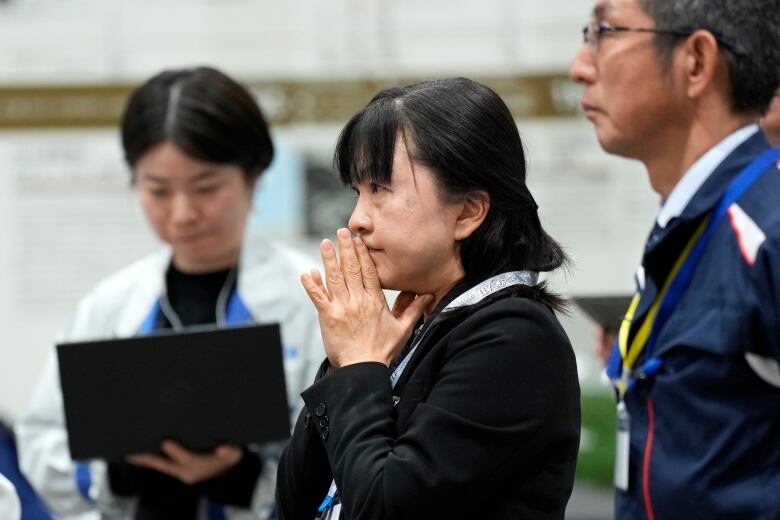 A woman clasps hands and has a look of concern on her face while standing. A man and a woman are seen in her proximity.