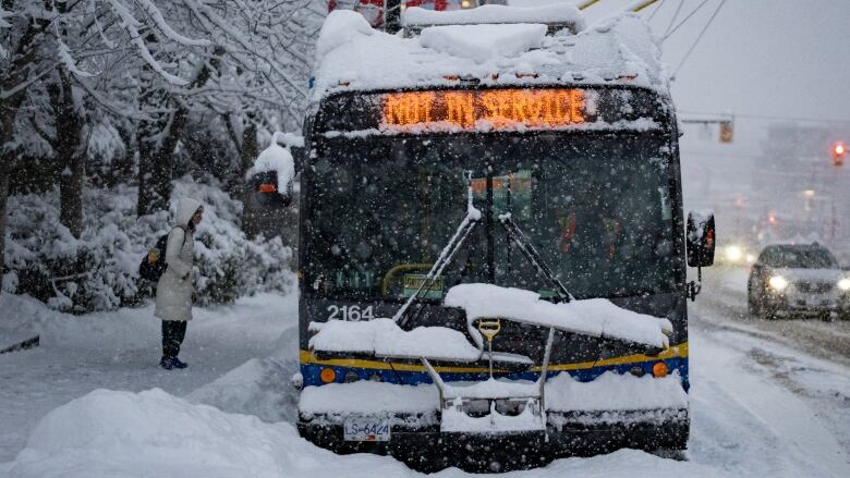 A person wearing a cream-coloured jacket waits outside a bus blanketed with snow amid a snowstorm. The bus chyron reads 'Out of service'.