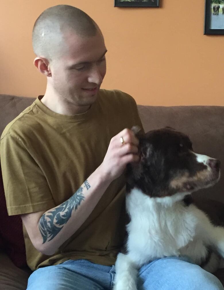 A slim man with large forearm tattoos and a shaved head pets a dog sitting in his lap.