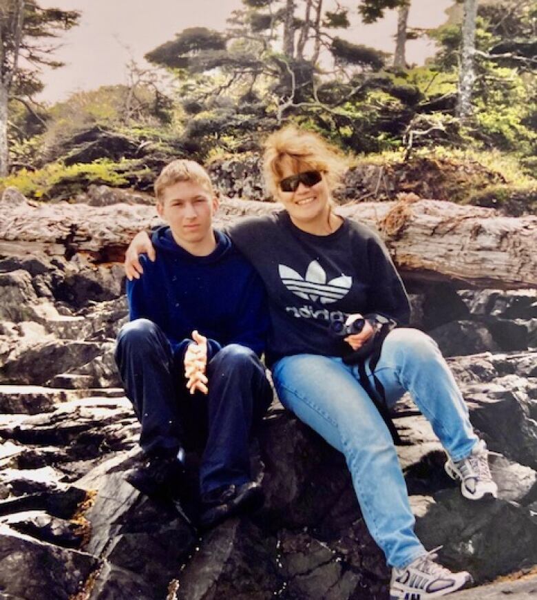 A teenage boy and a woman pose for a photo while sitting on rocks with trees in the background. 