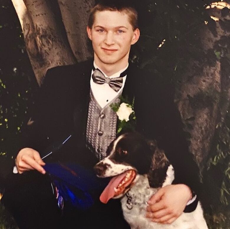 A young man in a tuxedo, kneeling in front of a tree, with his arm around a dog.