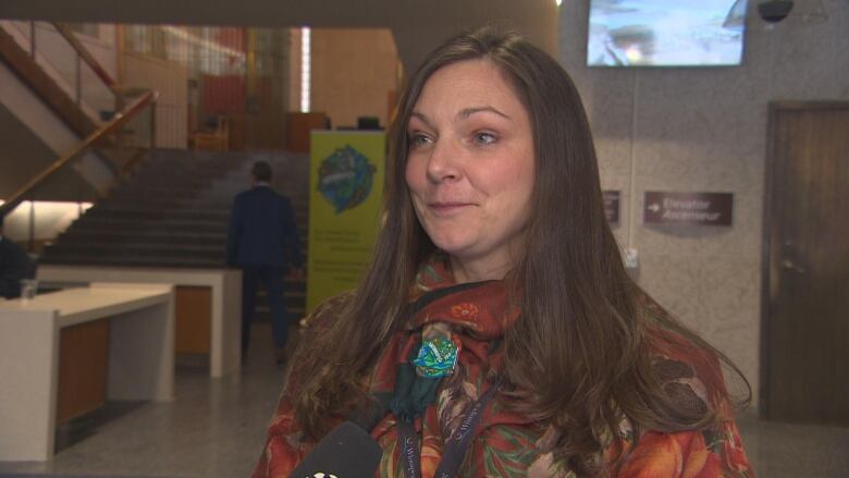 A woman with long brown hair in an orange-patterned shirt speaks into a reporter's microphone.