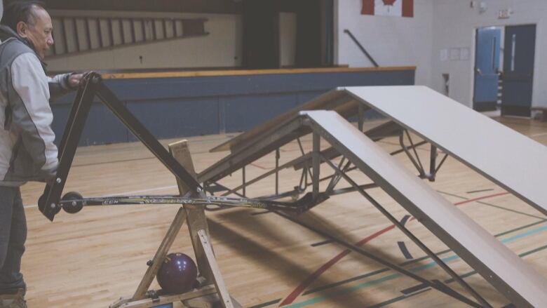 A man pushes down on a lever attached to two hockey sticks, which are lifting a folding table and folding it from the centre.