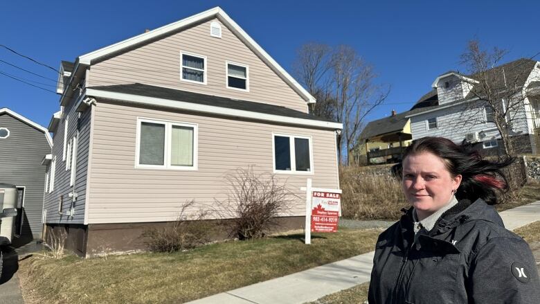 A woman with brown hair wearing a dark jacket stands in front of a two-storey home with a for sale sign in front.