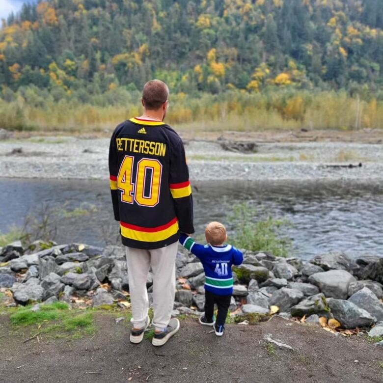 A father and tiny son wearing hockey jerseys look out at a river.