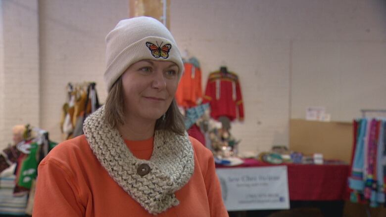 A middle-aged woman wearing a white beanie with a monarch buttery and a bright orange crewneck sweater. She has a beige woven scarf around her neck. 