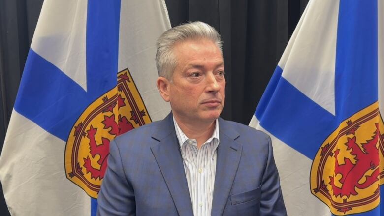 A man in a suit stands in front of two Nova Scotia flags.